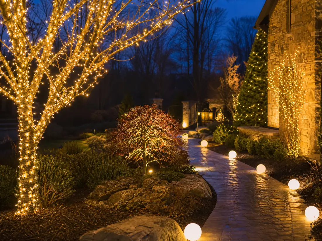 Warm-white wrapped trees and lit walkway globes lining a stone-home pathway at night